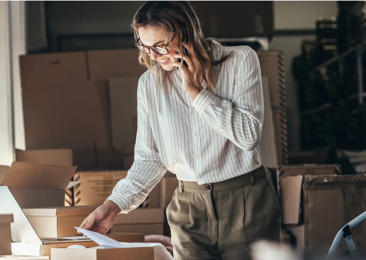 Smiling woman speaking on the phone while checking paperwork in a warehouse.